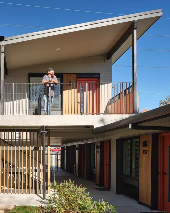 Man stands on an upper balcony of a modern apartment building with sloped roofs, red doors, and a clear blue sky.