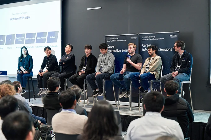 A group of individuals are seated on a stage, participating in a panel discussion. They hold microphones and appear engaged with an audience in front of them. Behind them, two screens display the words "Career Information Sessions" and "Reverse Interview," along with the logo for Hyundai Motor Group. Audience members are sitting and listening attentively.