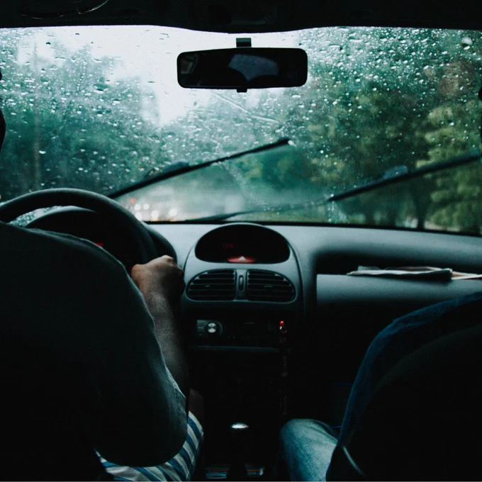 Image of a person driving a car on a rainy day, as seen from the backseat. Rain is visible on the windshield, with wipers actively clearing the view. The road ahead is blurred by rainfall.