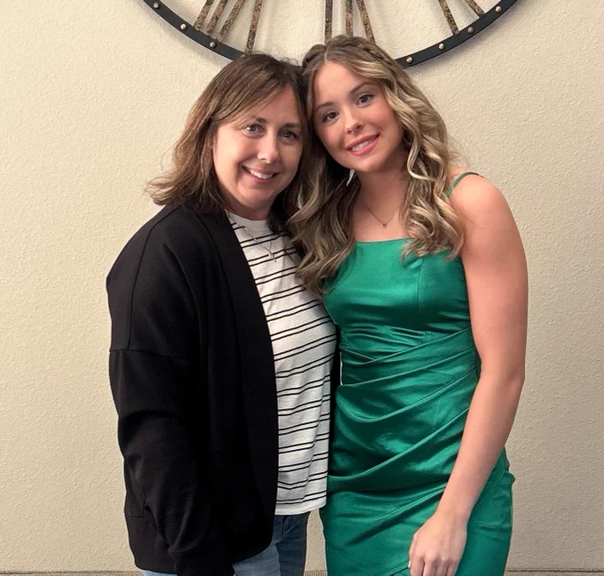 Two people stand in front of a wall with a decorative clock. They are smiling. One is dressed in a formal green dress.