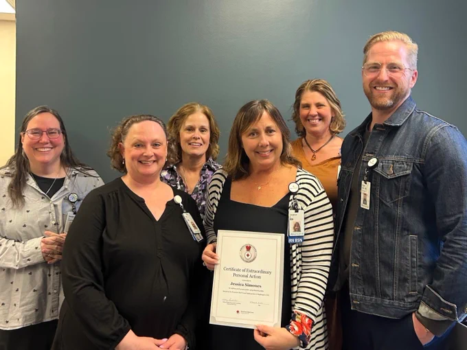 A group of six people smile while posing for a photo indoors. One person stands at the front holding a "Certificate of Extraordinary Personal Action" from the American Red Cross. The others, standing behind, are also smiling. All are wearing hospital badges.