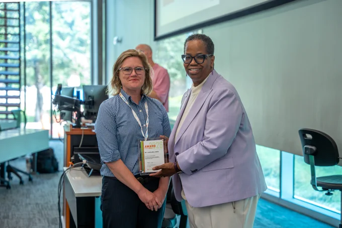 Dr. Caroline Jones, left, and Dean Adams, right, stand indoors; Dean Adams is handing an award plaque to the other, both facing the camera and smiling. classroom and computer equipment are visible in the background.