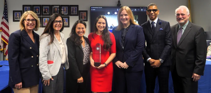 Photograph of seven professionally dressed individuals standing in a row indoors, with one person in the center holding a clear award or trophy. Background includes framed portraits on a wall, an American flag, and a screen displaying a virtual meeting.