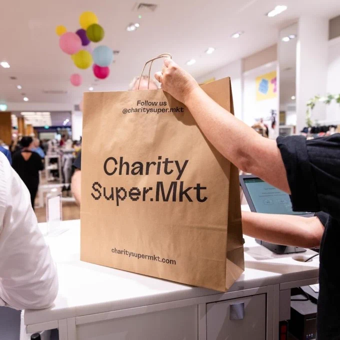 A person holds a brown shopping bag labelled "Charity Super.Mkt" at a store checkout counter. The store interior is bright, with colourful balloons in the background.