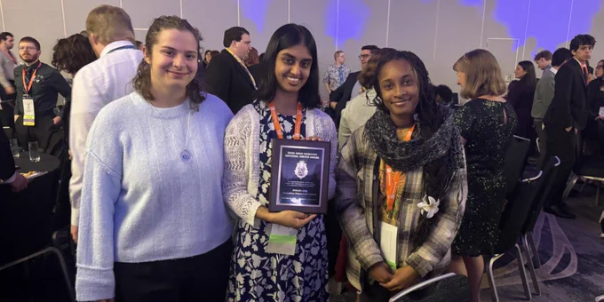 A person in the center holds a plaque at the Alpha Phi Omega National Convention. They stand with two individuals, all smiling. The event room is filled with people dressed formally.