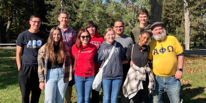 A group of ten people standing outdoors, smiling, in casual attire. Some are wearing shirts with Greek letters. There are trees in the background on a sunny day.