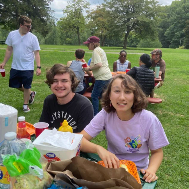 A group of people enjoy a picnic in a park. Two individuals sit at a table with snacks and condiments. In the background, others sit and stand near another table. Trees and a grassy area are visible.