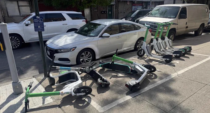 Sidewalk scene with several parked electric scooters lying haphazardly near a white Honda vehicle and a white van. A signpost and tree are visible in the background.
