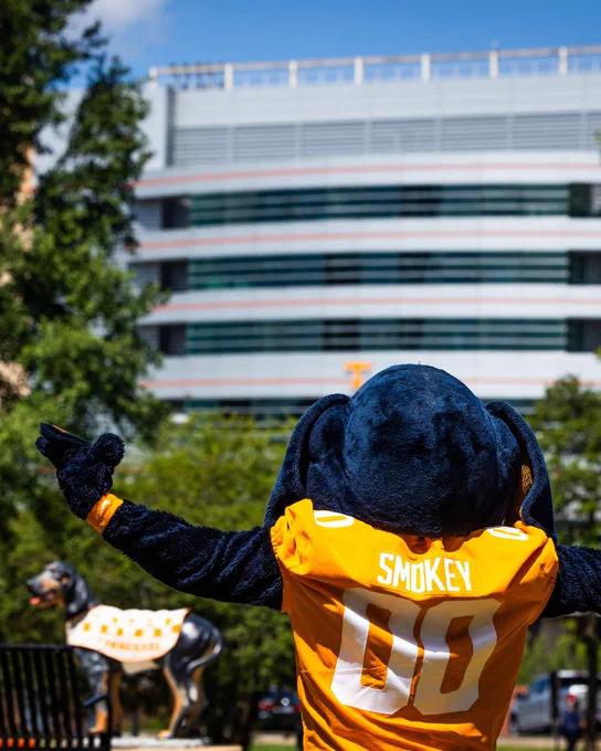 Smokey wearing an orange jersey with "Smokey" and the number "00" on the back stands with arms raised. In the background are trees and a multi-story building. A statue of Smokey featuring the Tennessee Volunteers theme is also visible.