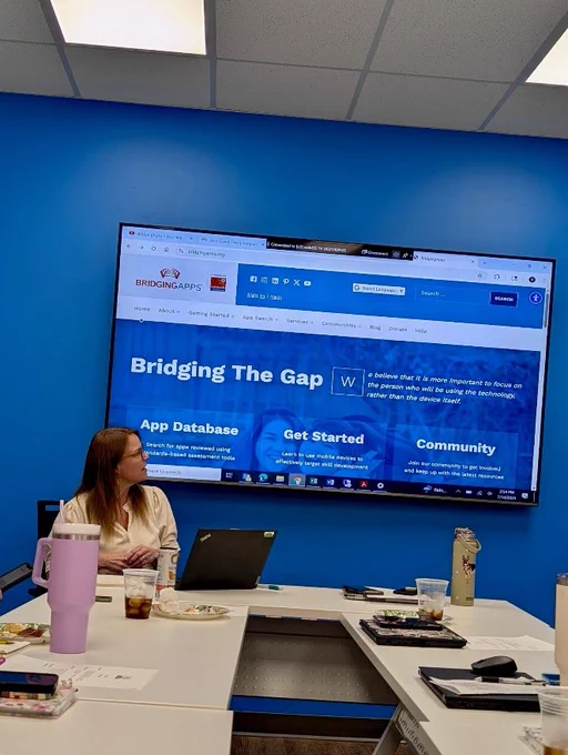 A person seated at a table with drinks and papers is looking at a large wall-mounted screen displaying a website titled "Bridging The Gap." The room has bright blue walls, and there's a laptop open on the table.