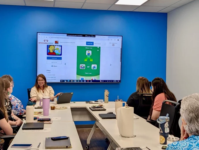 A group of people sit around a conference table in a room with bright blue walls. They are looking at a large screen displaying a website for Bridge and Apps. Various items like notebooks and water bottles are on the table.