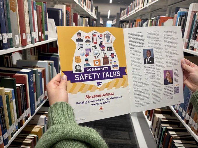 The image shows a person holding a booklet titled "Community Safety Talks" in a library aisle, surrounded by books on both sides. The booklet cover features illustrations related to safety, and an open page shows text and photos of presenters.