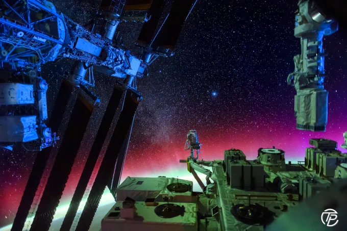 Vue de la Station spatiale internationale sous un ciel extrêmement étoilé, avec plusieurs structures et panneaux solaires visibles au premier plan. À l’horizon terrestre, des aurores polaires vertes et roses illuminent l’atmosphère en dégradés lumineux.