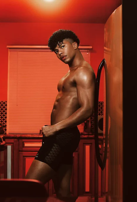 Muscular fit men, back side leaning against the fridge topless side looking at the camera with red kitchen background.