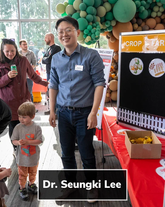 An indoor community science event with tables and balloon decorations in green and tan tones. An adult stands behind a red‑covered table with an educational display board showing crop and farm graphics. Several adults and a child stand nearby. A black text banner over the image reads, “Dr. Seungki Lee.”