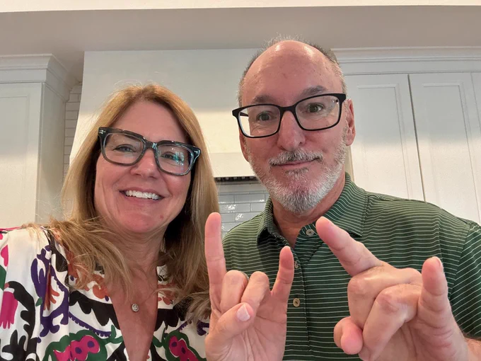Elizabeth Kyrstyn and Rick Fueyo smiling at the camera. They are both wearing black frame glasses and making "Horns Up" signs with their hands.