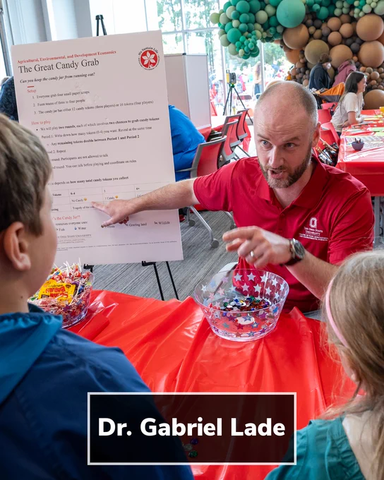 An indoor educational activity at a community event. An adult in a red shirt sits at a table covered with a red tablecloth, gesturing while explaining a game to children seated nearby. Clear bowls filled with colorful candy tokens are on the table. A sign on an easel behind the table reads, “The Great Candy Grab,” with instructions visible. Balloon decorations are in the background. A black text banner over the image reads, “Dr. Gabriel Lade.”