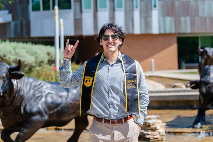 Evan Fueyo standing outdoorside the MSC near the bull statues at the University of South Florida. They are wearing a patterned shirt, beige pants, sunglasses, and a black graduation stole. They are smiling and making a "Horns Up" hand gesture. The background includes a building and greenery.