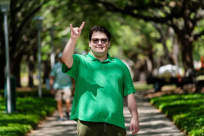 Jonathan Fueyo wearing a green polo shirt and sunglasses smiling and making a "Horns Up" hand sign. In the background is a path with greenery and large tress on both sides.