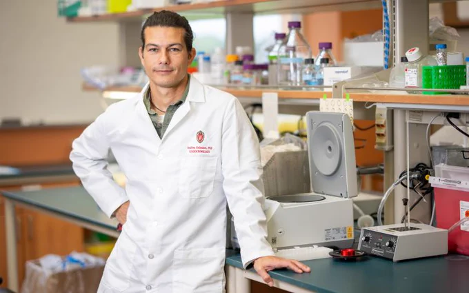 Scientist in a white lab coat standing confidently in a busy laboratory filled with scientific equipment and supplies.