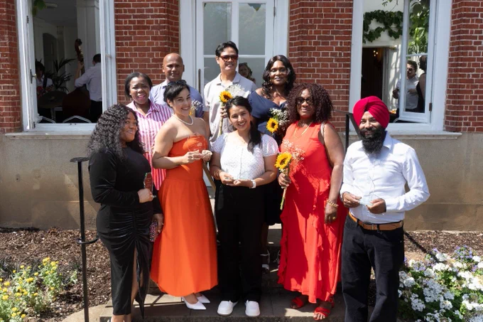 A diverse group of nine people, some holding trophies and sunflowers, pose for a photo outside York House.
