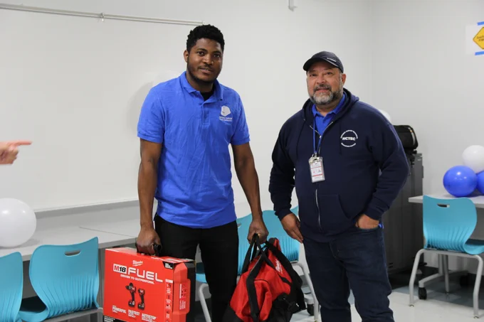 Two men standing indoors, one holding a red Milwaukee tool box and a black and red bag, the other wearing a navy jacket and cap.