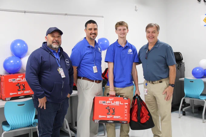 Four men standing in a classroom holding a red tool box and a black and red bag, with blue balloons in the background.