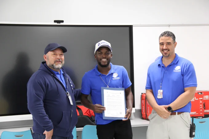 Three men in blue uniforms stand in front of a large screen, one holding a certificate in a classroom setting.