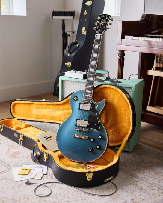 An antique pelham blue Gibson 1968 Les Paul Custom electric guitar resting inside an open hardshell case with a yellow interior. The case lies on a rug, surrounded by guitar-related items. In the background, there is an amplifier, another guitar case, and a piano.