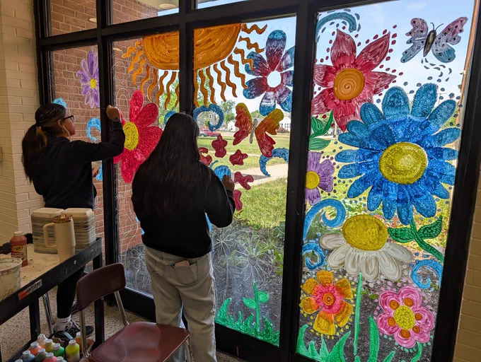 Two individuals are seen indoors, standing before floor-to-ceiling glass windows that face a sunny, grassy lawn with some buildings and cars in the distance. The windows are heavily decorated with colorful, painted murals depicting large flowers, a bright sun, a butterfly, and other swirling designs. One person on the left, with dark hair tied back and wearing a black hoodie and light pants, is positioned near a utility cart stocked with paint supplies, and is actively painting a purple and pink flower with a paintbrush. The second person, slightly taller, is positioned to their right, also with dark hair and wearing a black long-sleeved shirt, facing away from the camera and towards the windows, appearing to be observing or planning their next artistic addition.