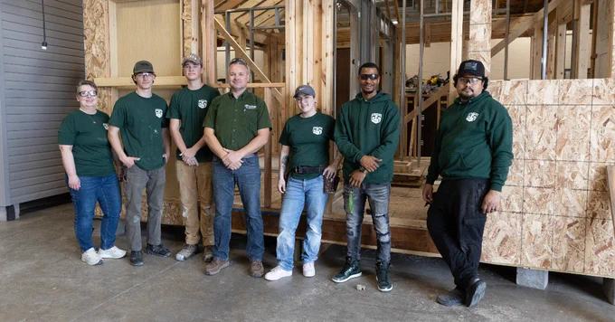 A group of seven individuals wearing green shirts stand indoors in front of a partially constructed wooden structure.