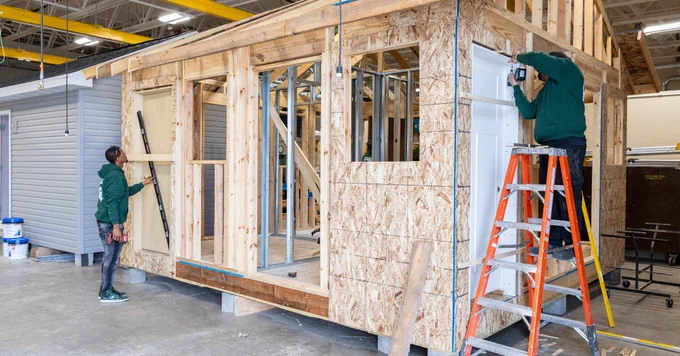 Two individuals are constructing a wooden framework of a house inside a large warehouse. 