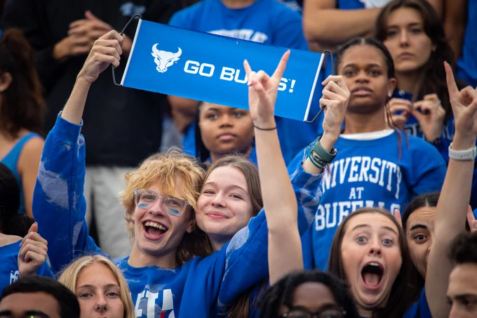 A crowd of University at Buffalo students, dressed in blue, cheers. One holds a \"GO BULLS!\" sign with the UB Bulls logo. The group shows excitement, with some smiling and others with their mouths open.