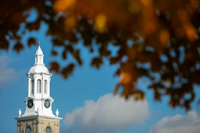A low angle captures a clock tower, its white architecture standing out against a bright blue sky with scattered clouds and out-of-focus autumn leaves.