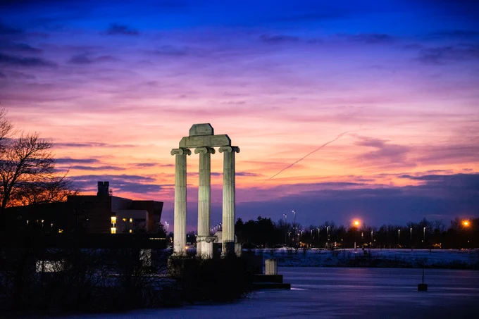 Twilight scene featuring an Ionic-style monument with three columns. A body of water in the foreground, with buildings and lights visible in the distance under a colorful sky. A vapor trail cuts through the clouds.