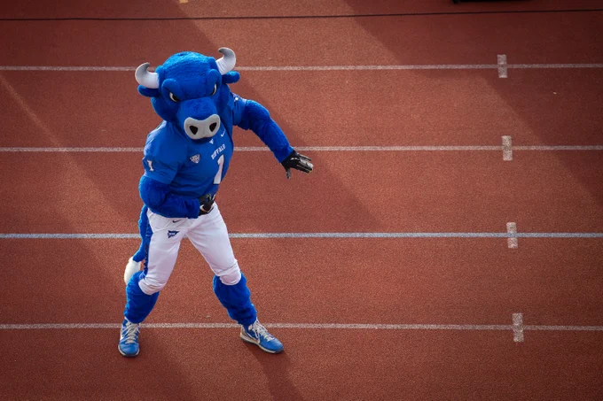 Victor E. Bull, the mascot with white pants and blue shoes, stands on a track with white lane markings. The mascot wears a blue jersey with the word \"BUFFALO\" and the number 1 on it.