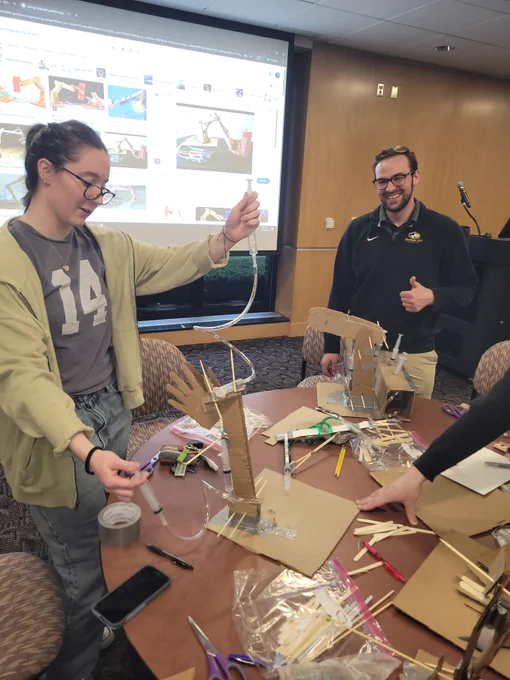 Two individuals working on a cardboard and popsicle stick engineering project in a classroom setting with tools and materials scattered on the table.