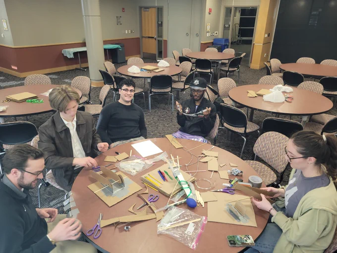 Five people seated around a table crafting with cardboard, scissors, and markers in a community room with round tables and chairs.