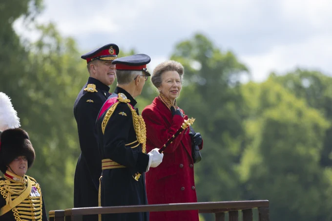HRH The Princess Royal and a group of people in formal military attire stand on an outdoor  platform. The individuals are wearing elaborate uniforms with gold accents. The setting appears to be a formal event, with trees visible in the background.
