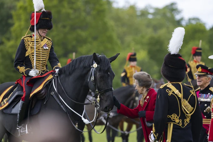 HRH The Princess Royal smiles and interacts with a member of the King's Troop Royal Horse Artillery who is mounted on a black horse. Other members of the regiment, all in ceremonial dress, are visible in the background. They are in an outdoor setting with greenery.