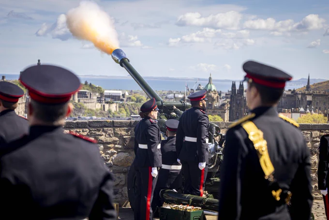 A group of uniformed individuals stands around a Light Artillery gun on a stone platform at Edinburgh Castle, while it fires a bright flame. The cityscape of Edinburgh, including historical buildings, is visible in the background under a partly cloudy sky.