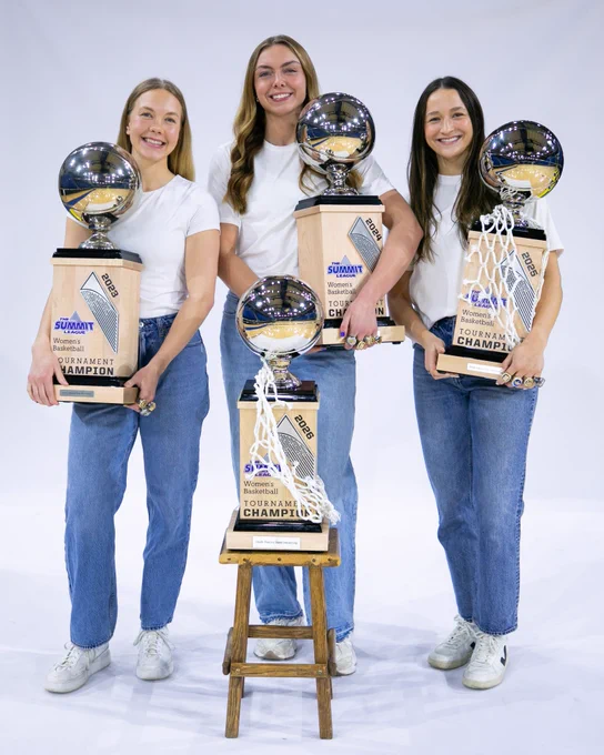 Studio portrait of three women's basketball players standing side by side against a plain white background, each wearing a white T-shirt, blue jeans and white shoes while holding large basketball championship trophies. A fourth trophy of the same style sits on a small wooden stool centered in front of the group. The plaques on the trophies include “Summit League Women’s Basketball Tournament Champions” text and year markings.