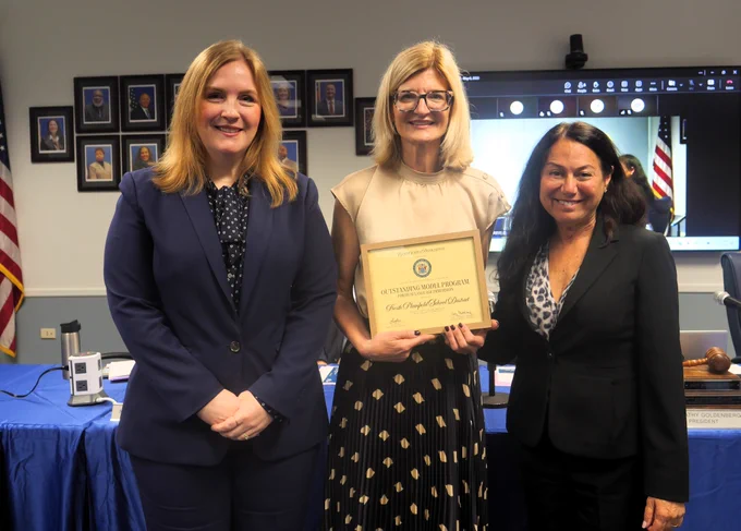 Photograph of three women standing indoors during an award presentation, with one woman holding a framed certificate. Background includes a blue tablecloth-covered table, framed portraits on a wall, and a screen displaying a virtual meeting with an American flag visible.
