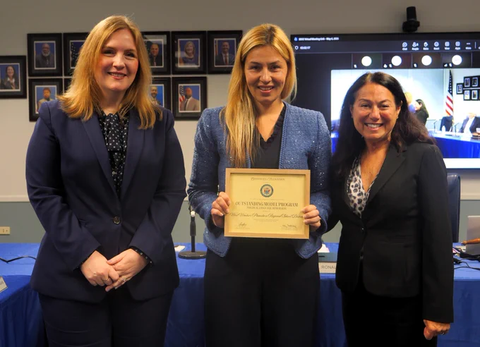 Photograph of three professionally dressed women standing indoors with one holding a certificate of recognition. Background includes framed portraits on a wall and a screen displaying a virtual meeting, indicating a formal award or recognition event.
