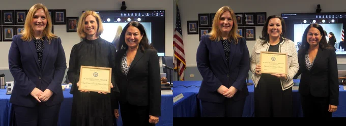 Photograph showing two separate groups of three women each standing indoors in front of a blue table and an American flag. Each group includes one woman holding a certificate, indicating an award or recognition event.