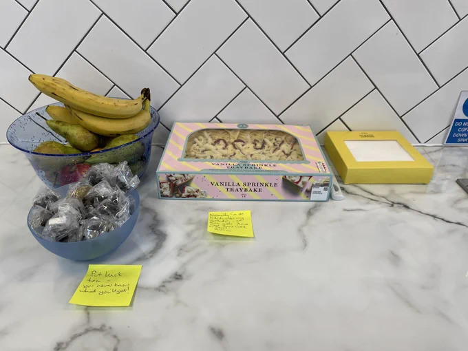 A work surface backed by white tiles with a vanilla sparkle traybake cake, a blue clear bowl with pears, apples and bananas and a small bowl with tea bags in small bags