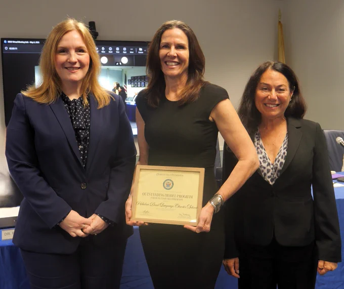 Photograph of three professionally dressed women standing indoors, with the center woman holding a framed certificate. Background includes a screen displaying a video call and a flag, indicating a formal or official setting such as an award ceremony or recognition event.
