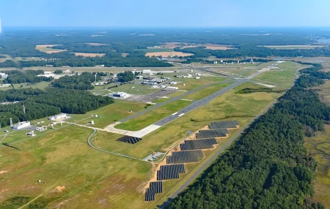 Aerial view of NASA's Wallops Flight Facility main base. Two aircraft runways with buildings scattered between surrounded by bright green grass. Below the runway in a field of grass is a solar panel farm along a long roadway and on the other side of the roadway is a dense forest of dark green trees.