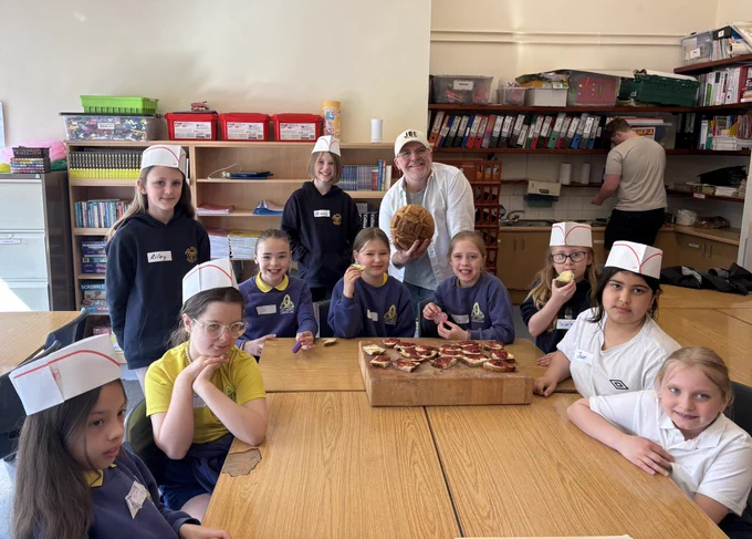 Group of children wearing paper chef hats gathered around a table with a wooden board holding mini pizzas in a classroom setting.
