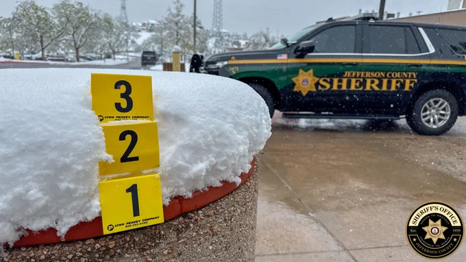 The image shows three yellow evidence markers labeled 1, 2, and 3 placed vertically in fresh snow on a concrete planter outside the Jefferson County Sheriff’s Office headquarters in Golden, Colorado. The snow reaches about halfway up marker 3. In the background, a Jefferson County Sheriff SUV is parked during active snowfall. The Sheriff’s Office logo appears in the bottom right corner of the image.